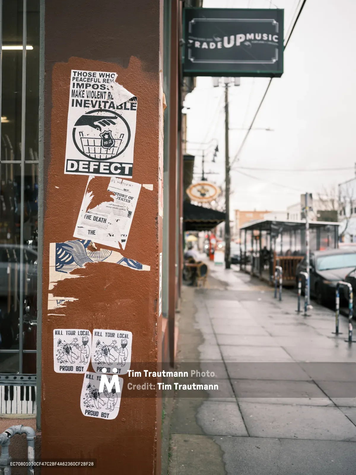 A weathered rust-colored utility pole on Northeast Alberta Street displays a collage of antifascist stickers and posters, including prominent messaging about peaceful revolution and defection. The urban scene captures Portland's activist street art culture against the backdrop of the Alberta Arts District, with Trade Up Music's storefront visible and wet pavement reflecting the overcast Pacific Northwest atmosphere. Layered political messaging creates a palimpsest of resistance culture on this busy commercial corridor.