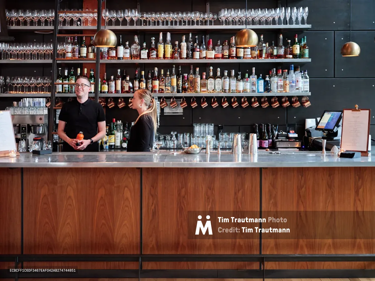 Two bartenders work behind a modern craft cocktail bar in Portland's Buckman neighborhood, surrounded by an impressive wall display of premium spirits and hanging glassware. The warm copper pendant lighting illuminates the rich walnut wood bar front, while crystalline wine glasses create geometric patterns on dark shelving above. The atmosphere captures the precision and artistry of contemporary cocktail culture in Oregon's vibrant hospitality scene.