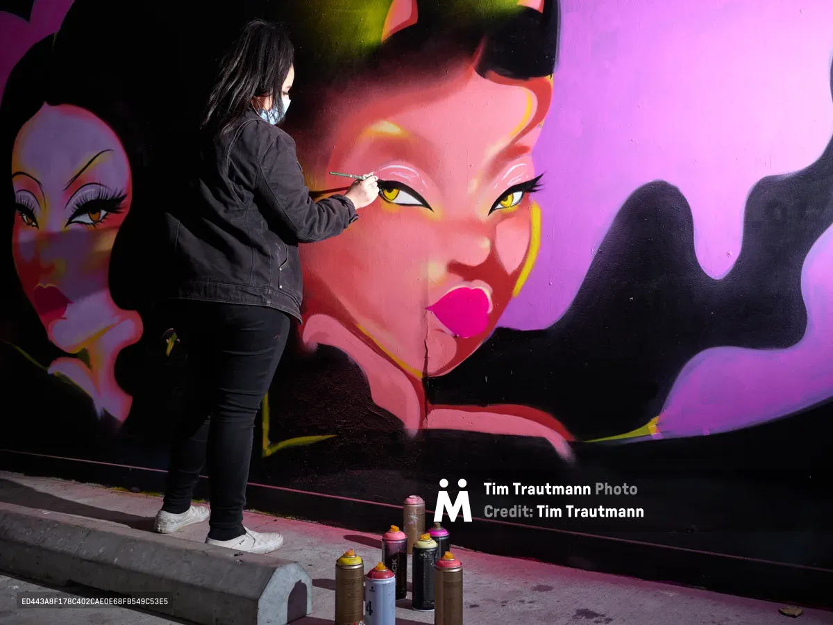 Multidisciplinary artist Vilatose meticulously applies detail to a striking mural featuring bold feminine faces rendered in electric pinks, purples, and yellows. Working in the intimate confines of Clay Creative's parking garage in Portland's Hosford-Abernethy neighborhood, the masked artist moves with practiced precision, surrounded by an arsenal of spray paint cans scattered across the concrete floor. The dramatic lighting illuminates both the artist's focused silhouette and the luminous portrait emerging on the wall, creating a compelling study of creative process in urban space.