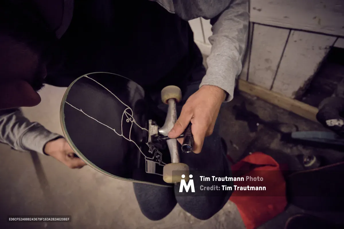 A skateboarder's weathered hands carefully adjust the trucks on a well-worn deck at Commonwealth Skateboarding in Portland's Buckman neighborhood. The moody lighting and intimate framing capture the ritual of board maintenance, with the skater's gray hoodie and striped sleeves creating visual texture against the dark skateboard. Red accents from clothing peek into the frame, adding warmth to the industrial setting of this Southeast Portland skate shop.