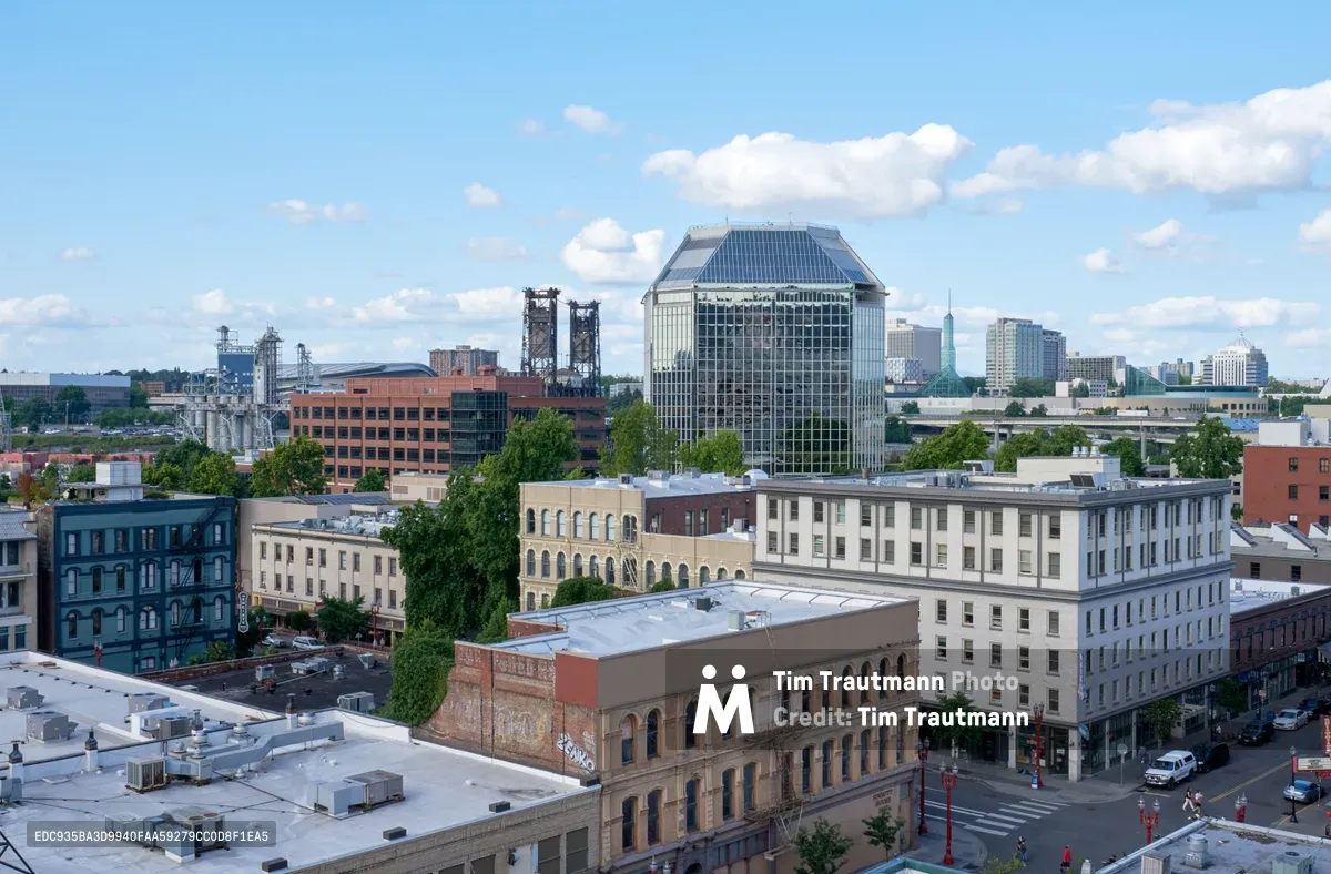 An elevated perspective captures Portland's historic Chinatown district in the foreground, where weathered brick buildings and converted warehouses create a layered urban tapestry. The distinctive glass tower of a major downtown building rises prominently in the middle distance, while the city's modern skyline stretches toward the horizon under a canvas of puffy white clouds against cerulean sky. Tree canopies punctuate the dense urban fabric, softening the architectural edges with verdant interruptions.