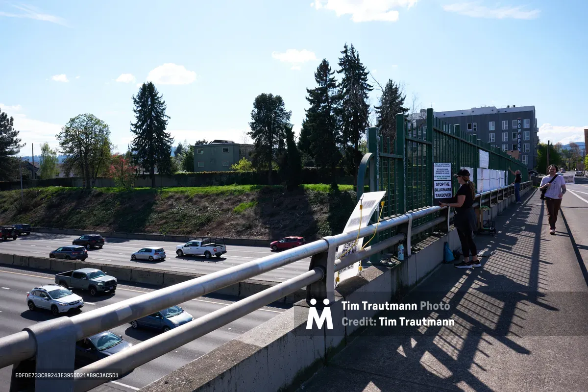 Beneath a clear Pacific Northwest sky dotted with white clouds, activists from the Indivisible movement unfurl a protest banner from a pedestrian overpass spanning Interstate 5 in North Portland. The concrete bridge frames a scene of urban activism, with participants silhouetted against the bright afternoon light as traffic flows steadily below. Towering evergreens and modern residential buildings create a distinctly Portland backdrop, while the activists' coordinated banner drop transforms this everyday infrastructure into a stage for political expression.