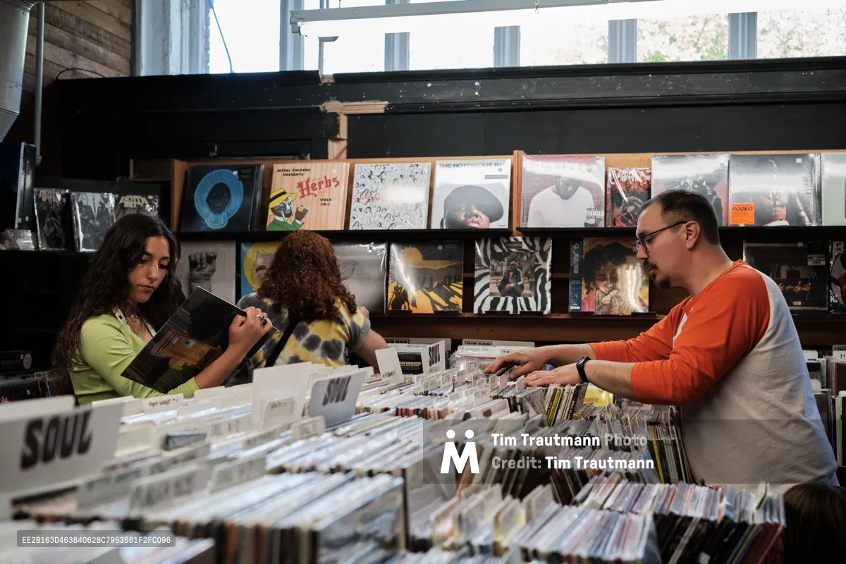 Three music enthusiasts browse through extensive vinyl collections at Music Millennium during the annual Record Store Day celebration in Portland's Kerns neighborhood. The woman in lime green examines album artwork while her curly-haired companion and a bearded man in orange systematically flip through alphabetized crates. Natural light filters through large windows, illuminating the store's carefully curated displays of classic and contemporary albums that line the walls above the browsing bins.