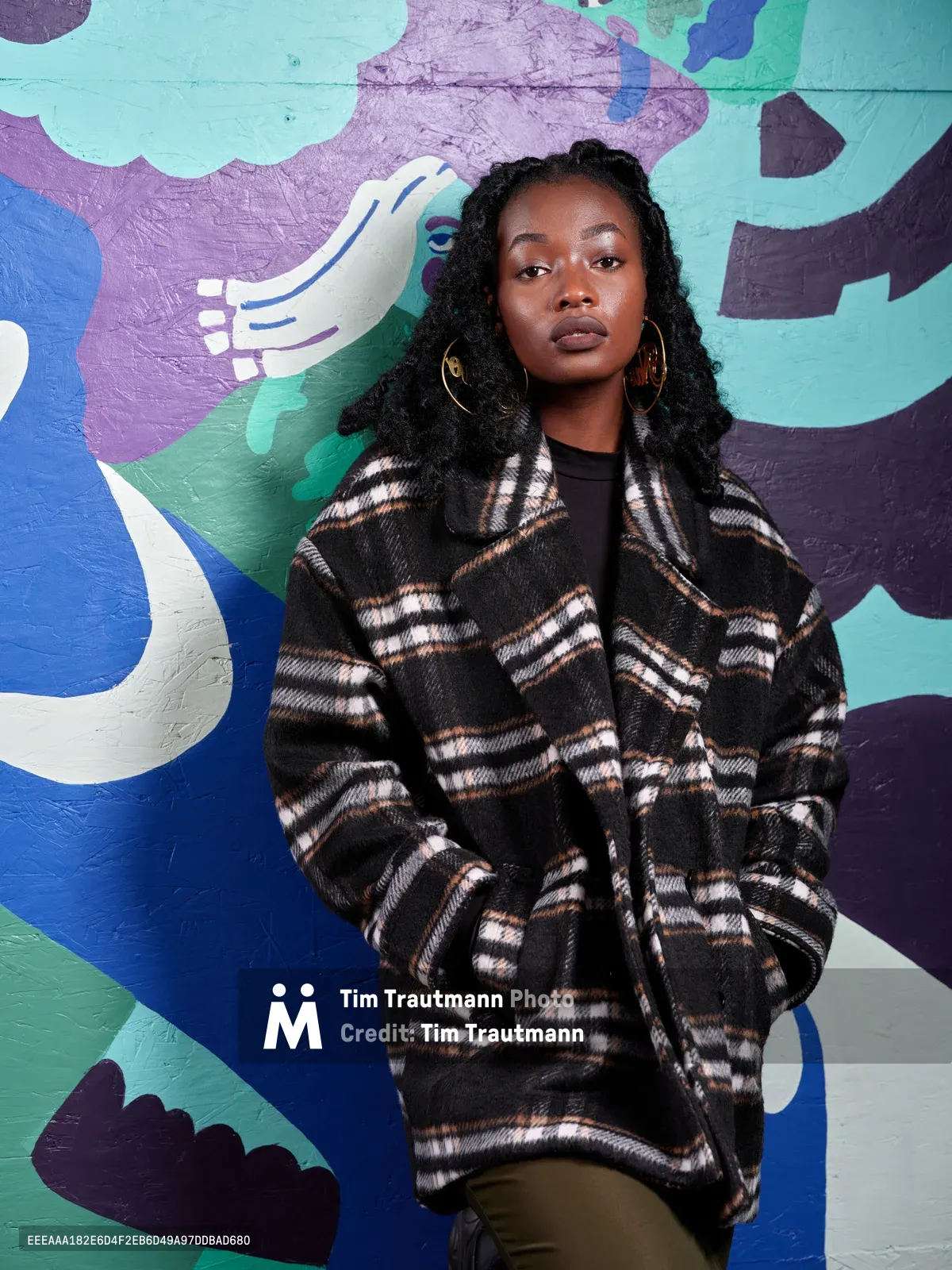 A portrait of artist Naomi Likayi standing against a vibrant painted mural in teal, blue, purple, and white. She wears an oversized black and white plaid wool coat over a black top, with olive trousers and large gold hoop earrings. Her long black braids frame her face as she gazes directly into the camera with a calm, composed expression.