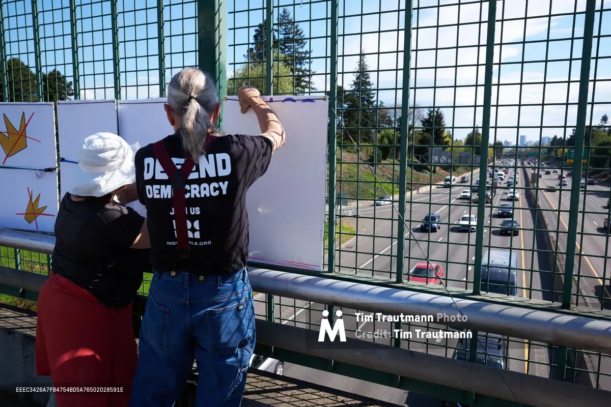 Two activists from the Indivisible movement position themselves on the North Skidmore Street overpass above Interstate 5 in Portland's Humboldt neighborhood, securing protest banners to the chain-link fence. The silver-haired man in a "Defend Democracy" t-shirt and his companion work methodically in the afternoon light, their white banner visible against the backdrop of moving traffic below. Evergreen trees frame the scene while cars flow steadily beneath this quiet act of civil engagement.