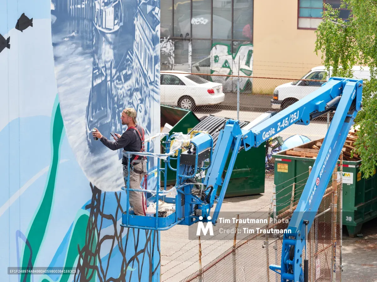 Jeremy Nichols and Devin Finley work methodically from a bright blue Genie aerial lift, applying paint to a sweeping mural on a building wall in Portland's Central Eastside district. The scene captures the industrial rhythm of street art creation, with construction debris and chain-link fencing framing the artists against their towering canvas of blues and greens. Afternoon light illuminates the working duo as they transform urban architecture into public art.