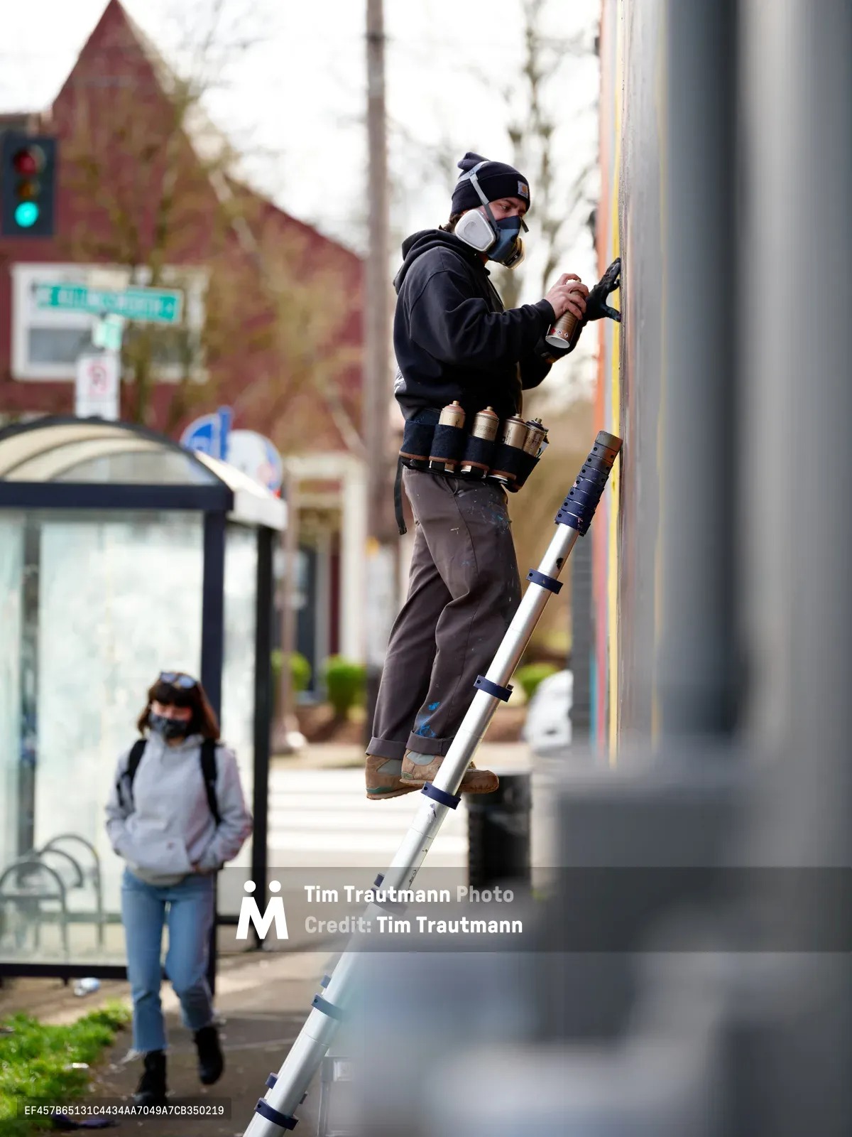 A masked street artist in dark clothing climbs an aluminum ladder to paint a colorful mural on a building wall along Northeast Killingsworth Street in Portland's Alberta Arts District. The urban scene captures the spirit of neighborhood revitalization, with a pedestrian in the background walking past a bus stop shelter. Soft daylight illuminates the artist's concentrated work, while the surrounding streetscape reveals the character of this gentrifying Portland neighborhood.