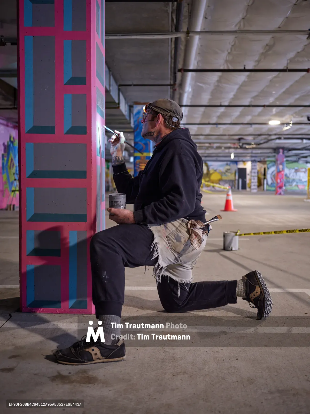 Portland artist and bike activist Rabbitt kneels in focused concentration as he paints his signature geometric pattern on a concrete column in the Clay Creative building's parking garage. The warm artificial lighting illuminates his work-worn hands applying pink and blue paint to create bold rectangular shapes, while the industrial space around him buzzes with creative energy. Paint cans and brushes scattered at his feet speak to the methodical process of transforming this utilitarian space into an artistic statement.