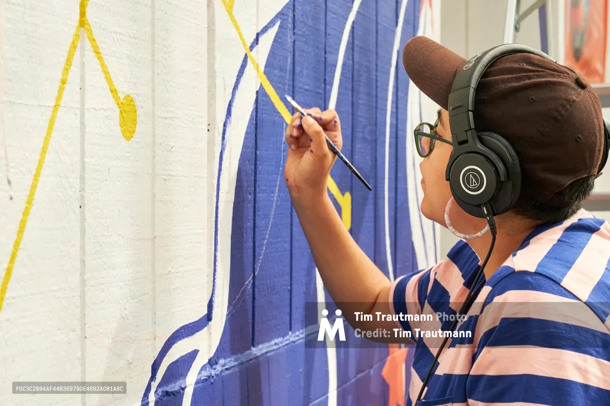 Maria Rodriguez, known as Sparkykneecap, applies vibrant blue and yellow paint to a wall at Open Signal in Portland's Eliot neighborhood. The Mexican-American artist wears headphones and a striped shirt while working on the commissioned mural titled "Let's Talk," their brush carefully adding flowing lines to the geometric composition. Warm afternoon light illuminates the creative process as Rodriguez transforms the white wall into a playful exploration of identity and community connection.