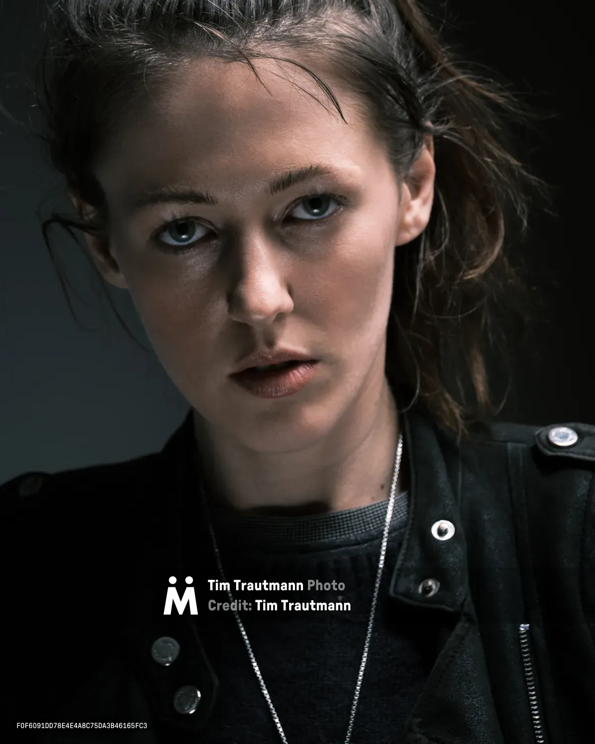 Rhianna Atwood gazes directly at the camera with piercing blue eyes in this intimate portrait captured in Portland's historic Irvington district. Tousled brown hair frames her face while she wears a black leather jacket over a dark mesh top, accented by a delicate silver chain necklace. The moody lighting creates dramatic shadows that emphasize the contemplative intensity of her expression against the neutral background.