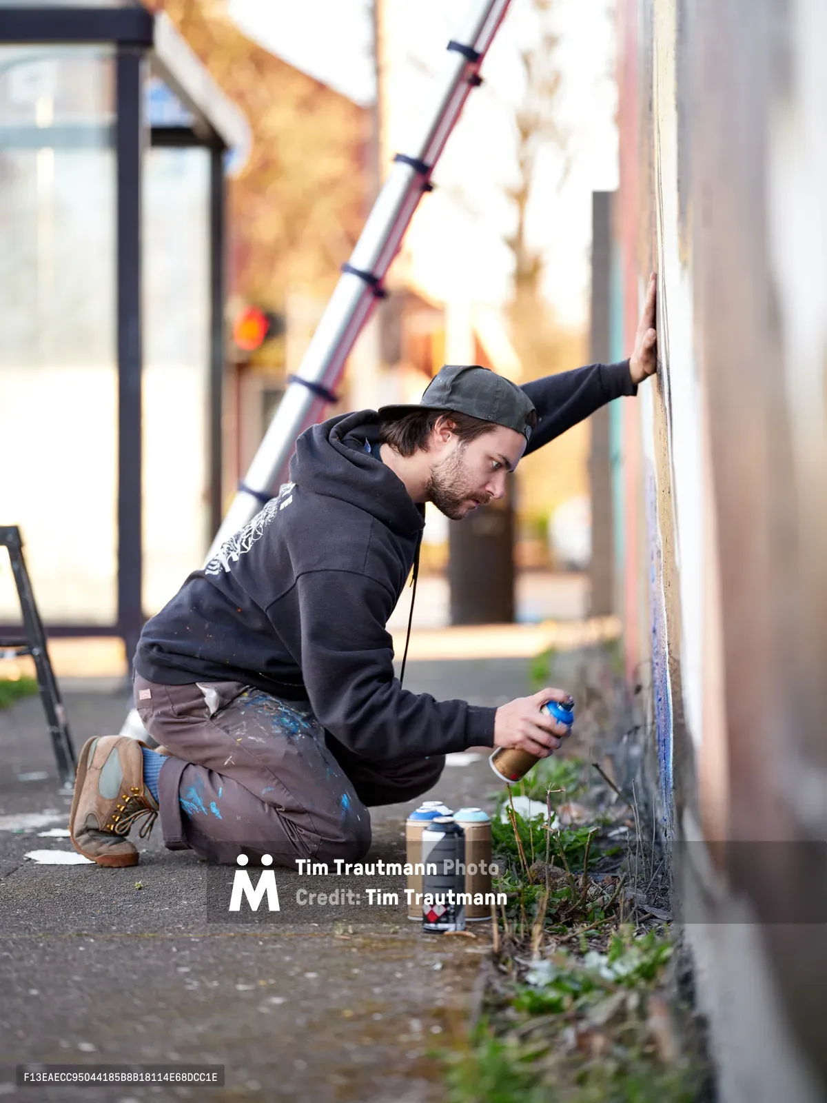 Travis Fields, known as campographic, crouches intensely beside a concrete wall on Northeast Killingsworth Street, spray paint can in hand as he works on a mural tribute to poet Amanda Gorman. The artist, dressed in paint-splattered clothing and a baseball cap, is captured mid-creation in the warm afternoon light of Portland's Alberta district. Scattered spray cans and small plants emerging from sidewalk cracks frame the intimate scene of artistic dedication.
