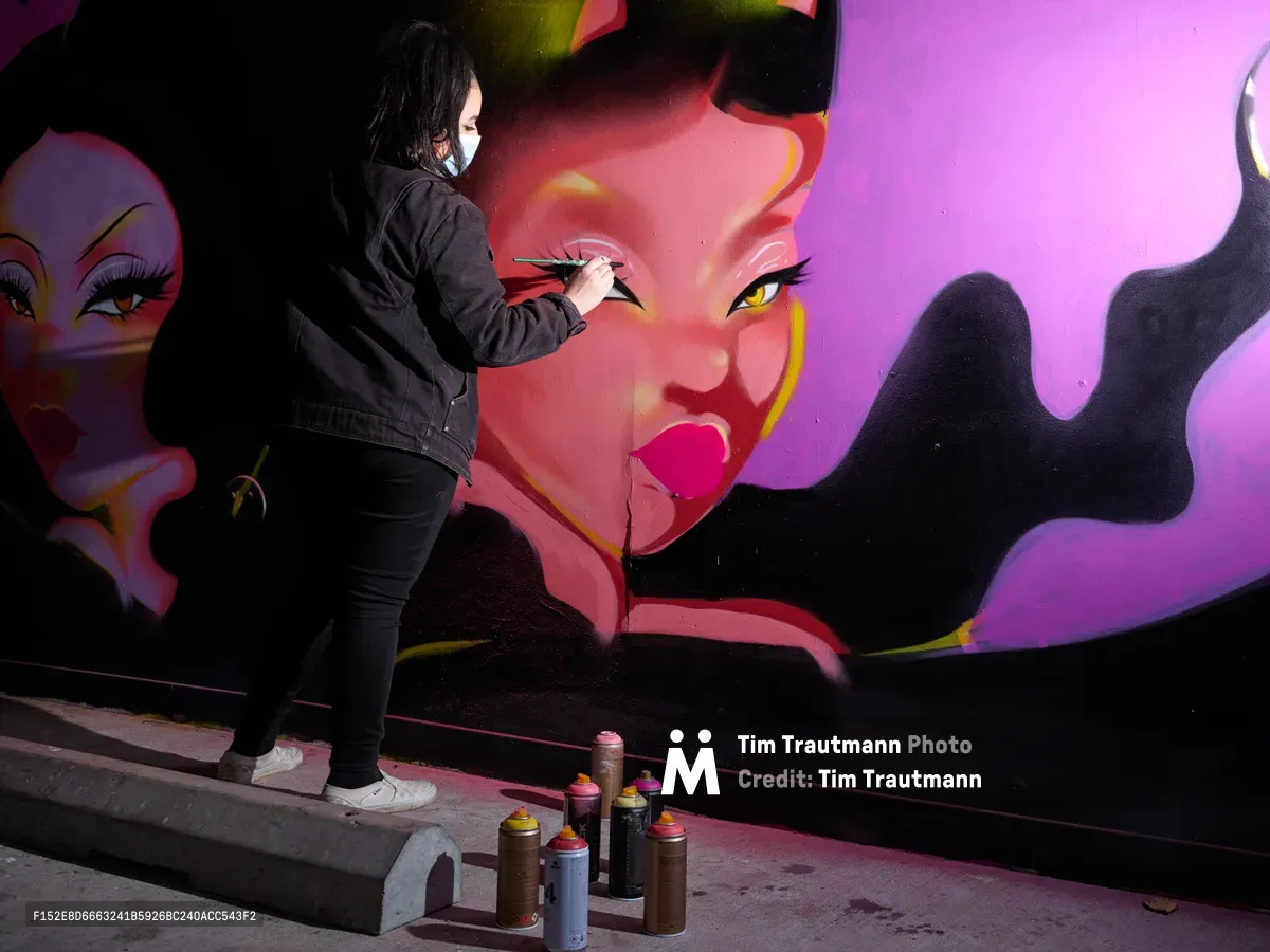 Multidisciplinary artist Vilatose meticulously details a striking magenta-toned portrait on the concrete wall of Clay Creative's parking garage in Portland's Hosford-Abernethy neighborhood. The masked artist, dressed in black, adds precise brushstrokes to luminous feminine faces that emerge from swirling purple and pink gradients. Scattered spray paint cans at her feet reflect the urban canvas transformed into gallery space, where raw industrial architecture meets contemporary street art.