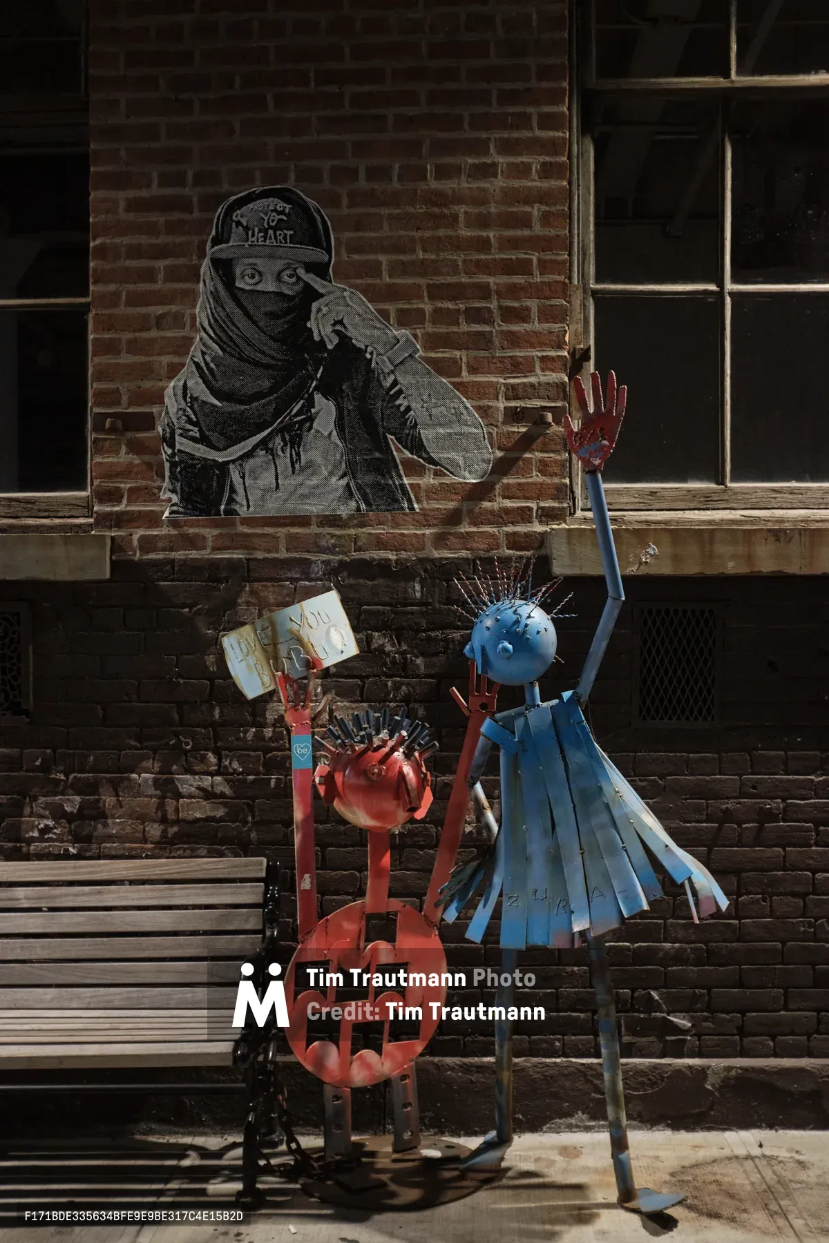 A striking urban tableau unfolds on Berry Street in Williamsburg, Brooklyn, where weathered brick walls frame a powerful dialogue between street art and sculptural installation. A monochromatic mural depicts a masked figure in contemplative pose, their piercing gaze watching over brightly painted metal sculptures below—a crimson jack-o'-lantern and azure figure with upraised arm create a vivid foreground against the industrial backdrop. Harsh daylight casts sharp shadows across the weathered sidewalk, emphasizing the raw texture of urban decay and creative resilience.