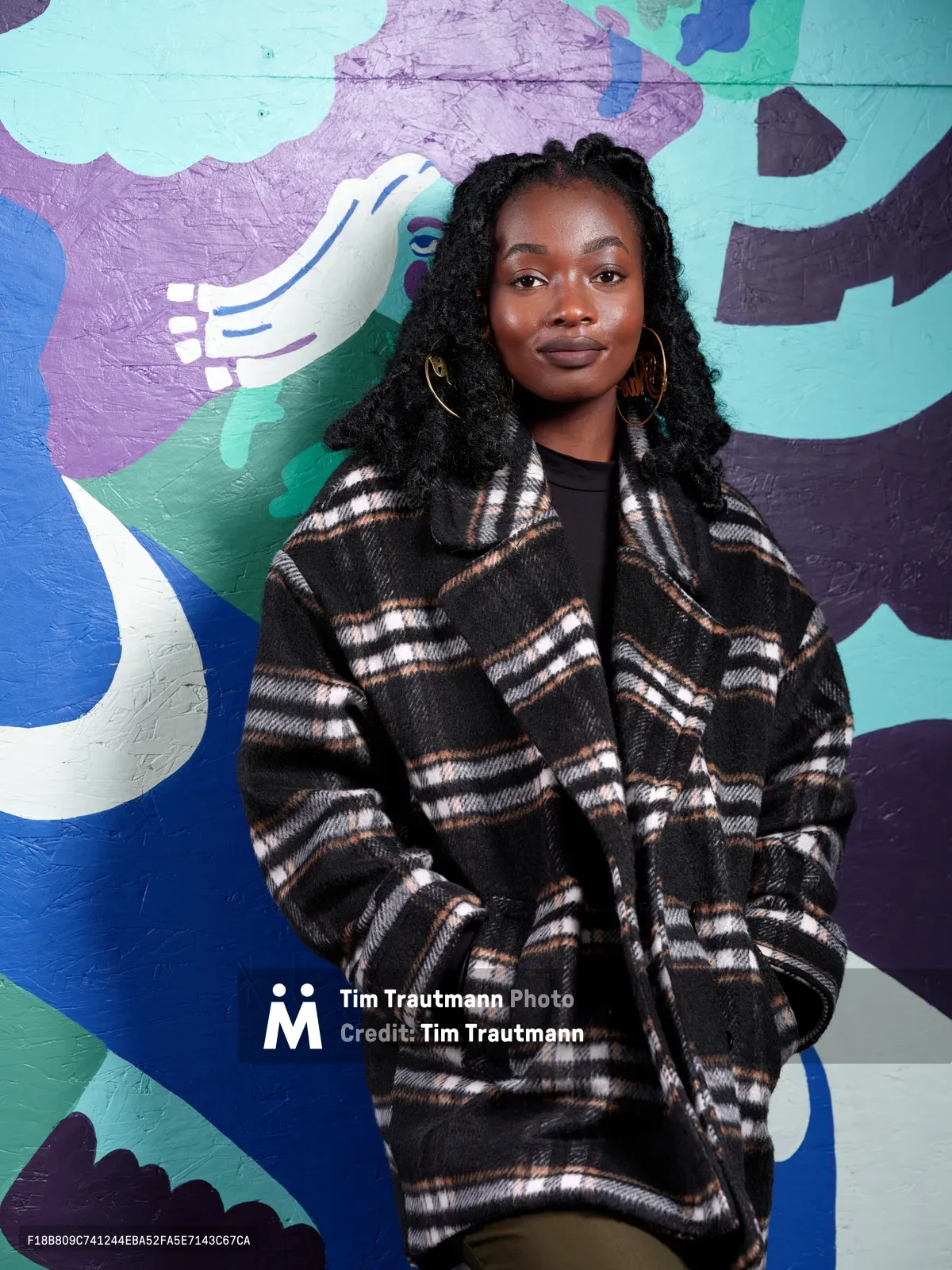 Artist Naomi Likayi stands confidently against her striking mural work on the boarded-up Portland World Trade Center, commissioned by the Portland Street Art Alliance. Her contemplative gaze and relaxed pose complement the sweeping abstract forms behind her—bold strokes of lavender, turquoise, and deep navy that seem to flow like organic shapes across the wall. The interplay between her textured plaid coat and the fluid painted forms creates a compelling dialogue between personal style and artistic expression in this urban canvas.