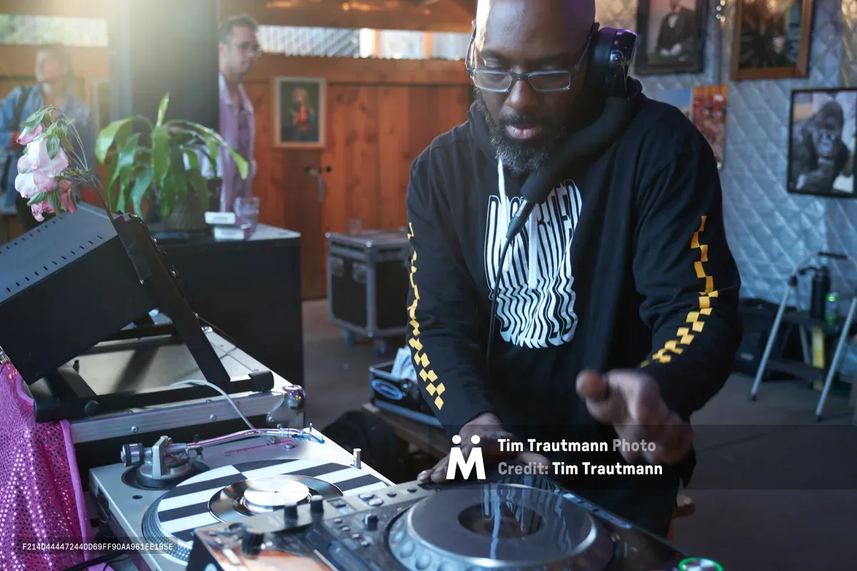 DJ Cee White commands the turntables during the Your Sunday Best season opener at White Owl Social Club in Portland's vibrant music scene. Warm afternoon light filters through the intimate venue as the bearded performer, wearing a black hoodie with yellow checkerboard details, manipulates vinyl records with practiced precision. The cozy club atmosphere, complete with potted plants and framed artwork on wood-paneled walls, creates the perfect backdrop for this daytime celebration of hip-hop culture.