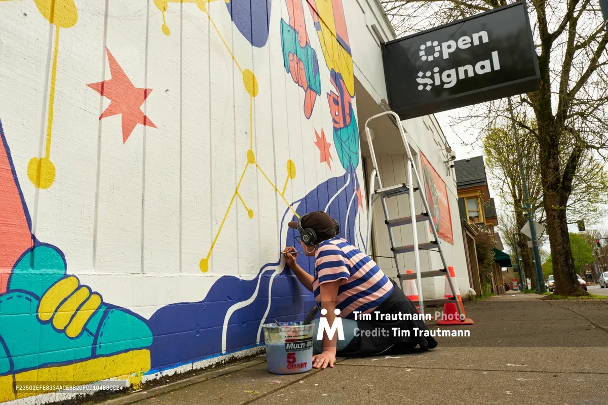 Mexican-American artist Maria Rodriguez, known as Sparkykneecap, kneels on the sidewalk while painting vibrant details on a colorful mural titled "Let's Talk" outside Open Signal in Portland's Eliot neighborhood. The artist, wearing a blue and white striped shirt, carefully applies paint to the geometric composition featuring turquoise, yellow, coral, and navy blue shapes against the white corrugated metal wall. A paint bucket sits nearby on the concrete as bare trees frame the community art project on Northeast Martin Luther King Jr. Boulevard.