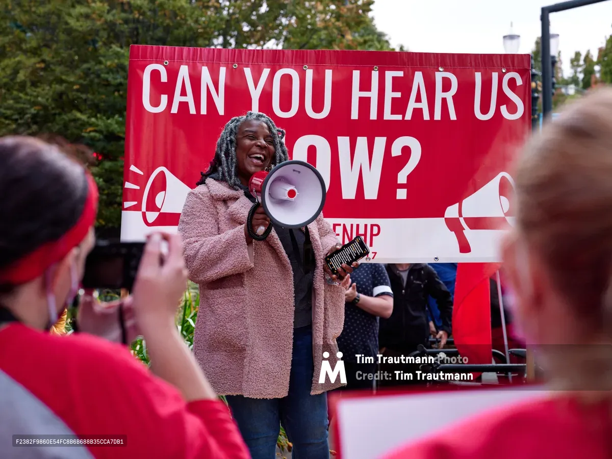A determined healthcare worker with dreadlocks commands attention through a red and white megaphone, her infectious smile contrasting with the serious nature of the Kaiser Permanente nurses' strike in Portland's Lloyd District. Behind her, a bold crimson banner emblazoned with "CAN YOU HEAR US NOW?" amplifies her message to the sea of red-clad supporters gathered in the foreground. The late afternoon light filters through verdant trees, casting a warm glow over this moment of collective action outside the towering Kaiser Permanente headquarters on Northeast Multnomah Street.