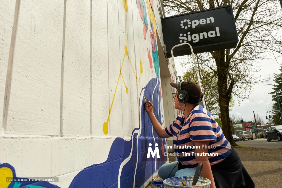 Maria Rodriguez, known as Sparkykneecap, meticulously applies blue paint to a vibrant mural on the white corrugated exterior of Open Signal in Portland's Eliot neighborhood. The Mexican-American artist, wearing headphones and a striped shirt, works from a ladder while creating the commissioned piece titled "Let's Talk" for the Regional Arts & Culture Council. Soft spring light filters through bare tree branches, illuminating the evolving artwork that explores themes of identity and cultural connection through bold geometric shapes and vivid colors.