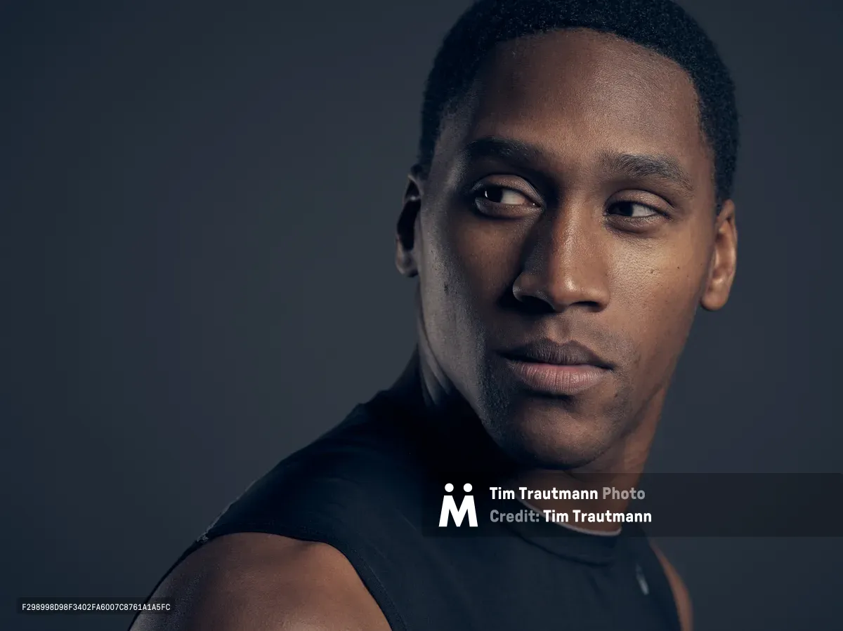 A striking studio portrait captures a young Black man in three-quarter profile against a moody charcoal background. Shot in Portland's Northwest District, the subject gazes thoughtfully off-camera while wearing a black crew neck shirt, his expression conveying quiet intensity and introspection. The dramatic lighting sculpts his features with precision, creating deep shadows that emphasize the contemplative mood of this professional headshot.