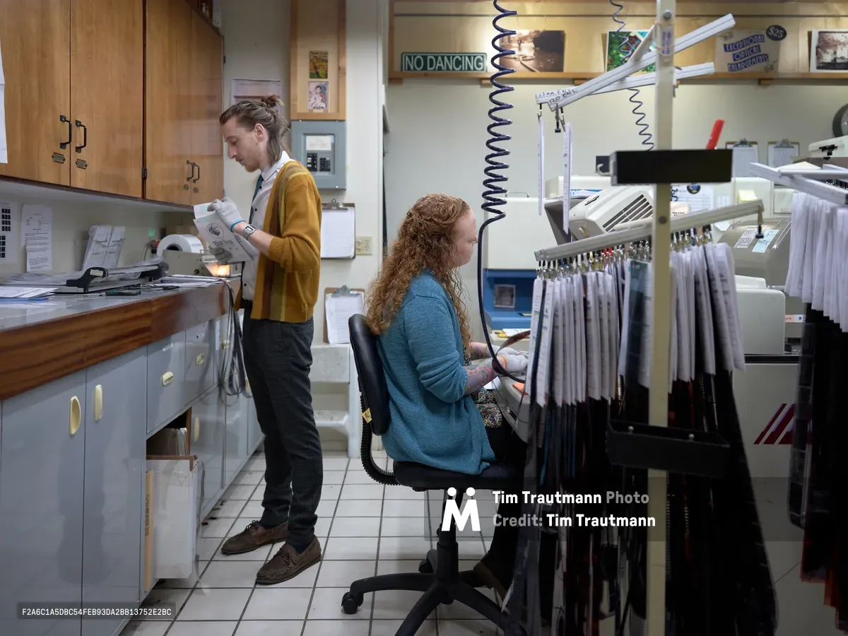 Two film lab technicians work in the intimate, equipment-filled processing room at Blue Moon Camera & Machine in Portland's Saint Johns neighborhood. A woman with curly red hair sits at a professional film processing station while her colleague in a mustard yellow cardigan examines negatives by the counter. The warm fluorescent lighting illuminates vintage processing equipment, coiled cords, and the organized chaos of a working analog photography lab.