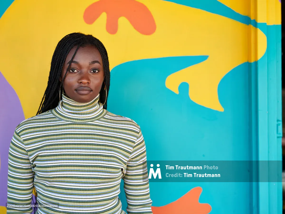 Naomi Likayi stands confidently before her completed mural in Portland's Alberta Arts District, her long braids framing her face as she poses in a striped turtleneck. The explosive backdrop features sweeping organic forms in brilliant turquoise, sunshine yellow, and coral orange that dance across the wall behind her. Natural light illuminates both artist and artwork, creating a harmonious dialogue between creator and creation in this intimate moment of artistic completion.