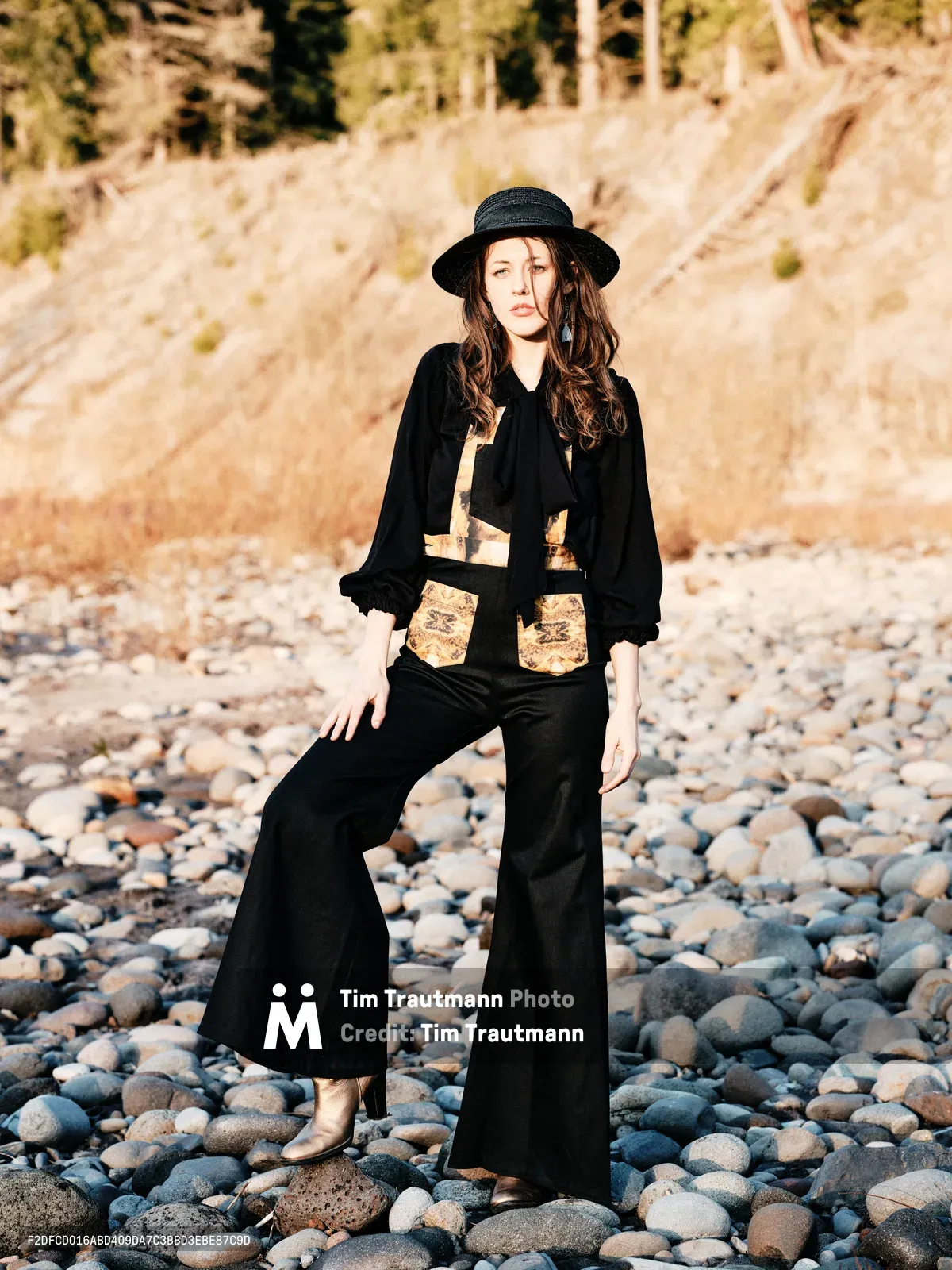 A young woman stands contemplatively on a riverbed of weathered stones along the Floodplain Trail in Multnomah County, Oregon. She wears a striking black ensemble with metallic embellishments and a wide-brimmed hat, her flowing hair catching the golden hour light. The composition captures her against the textured landscape of river rocks and distant hillsides, creating an atmospheric portrait that blends fashion with the raw beauty of the Pacific Northwest wilderness.