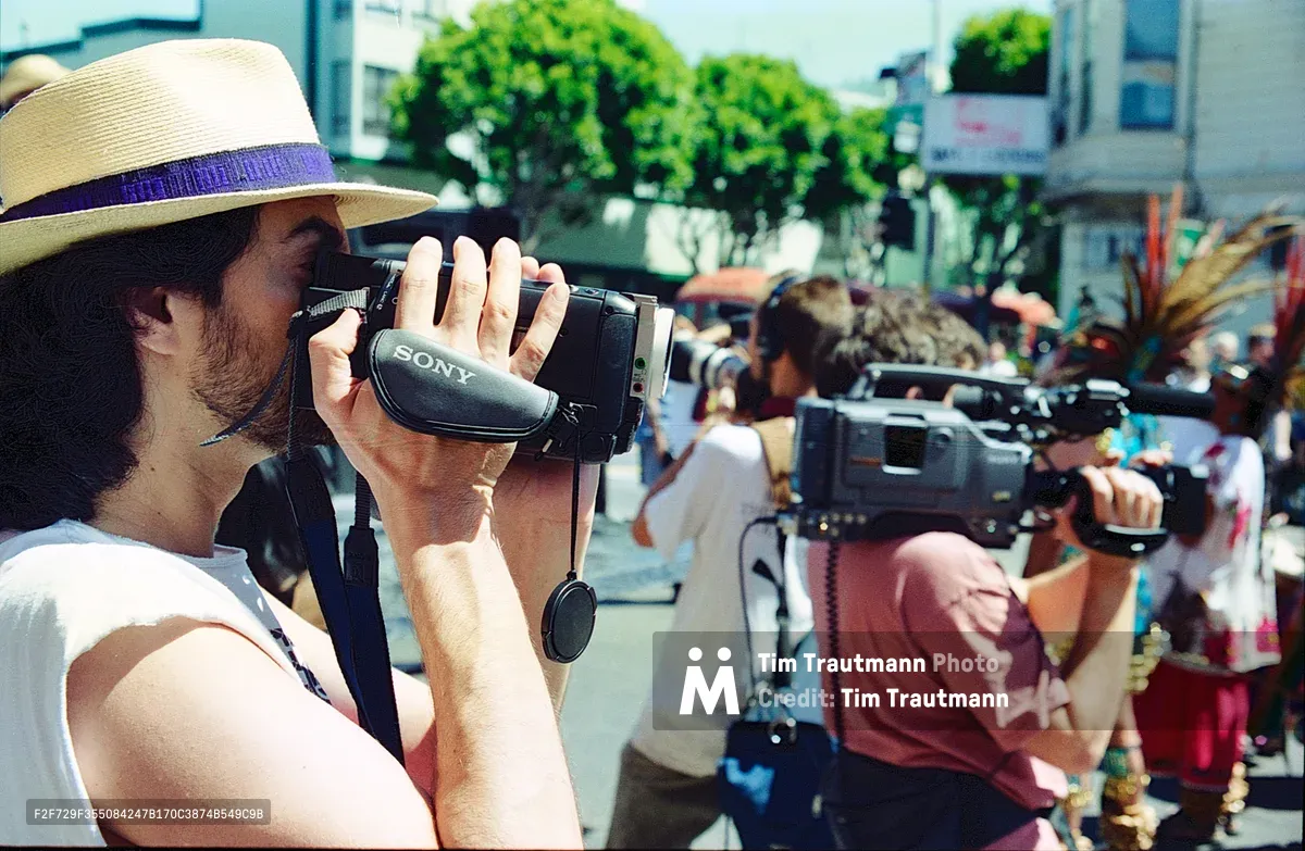 A bearded man in a woven straw hat with purple band operates a handheld Sony video camera in the foreground, while another person with blonde hair films behind him during a community demonstration. The scene unfolds on a sun-drenched Mission District street, with leafy green trees and urban buildings creating a backdrop of neighborhood activism. The warm afternoon light captures the grassroots nature of residents documenting their own struggle against gentrification.
