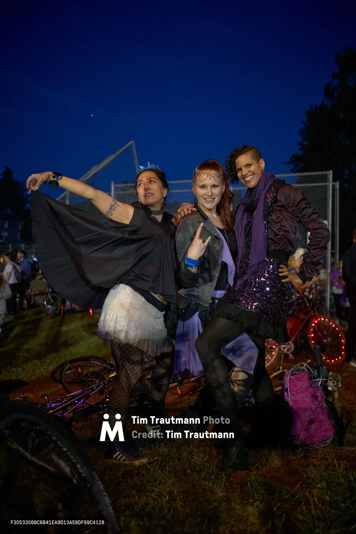 Three costumed participants pose exuberantly against the deepening cobalt sky at Portland's Drop Out Prom Ride during Pedalpalooza 2019. The woman on the left dramatically extends her black cape while balancing on decorated bicycles, her companion flashes a peace sign in purple attire and ornate headpiece, and their friend beams in sequined costume. Scattered props, LED lights, and fellow revelers create an enchanting tableau in Colonel Summers Community Garden as dusk settles over this unconventional celebration.