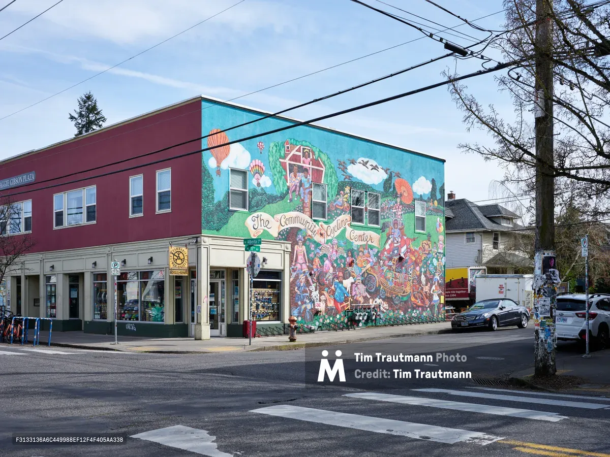 A vibrant community mural adorns the side wall of the Community Cycling Center on Northeast Alberta Street in Portland, Oregon. The whimsical artwork depicts hot air balloons, cyclists, and diverse community members in a celebration of local neighborhood life. Power lines cross the pale blue sky above the burgundy and white building, while parked cars line the quiet residential street under the soft overcast light of day.