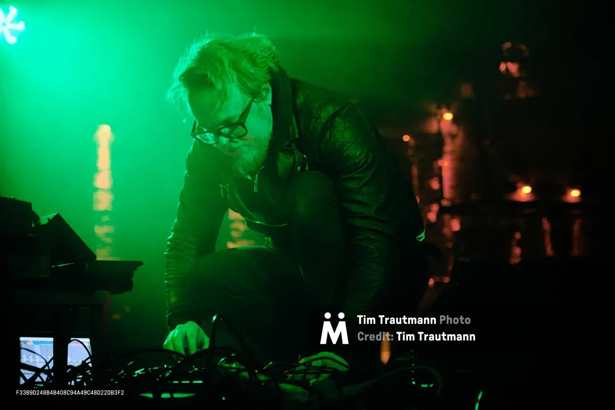 Electronic musician Eli Savage, performing as ellipsism, hunches intensely over his synthesizer setup in the emerald glow of stage lighting at Dante's in Portland. The atmospheric green wash transforms the intimate venue into an otherworldly sonic laboratory, while warm amber lights punctuate the darkness behind him. His focused posture and tousled hair capture the raw concentration of live electronic performance, as cables and equipment sprawl across the stage like digital arteries feeding his sonic creation.