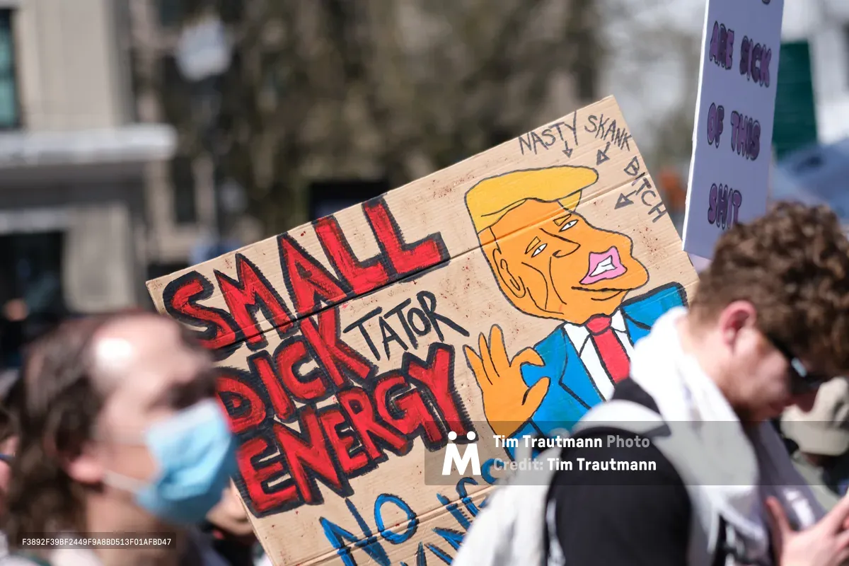 A handmade cardboard protest sign dominates the frame during a March 2026 demonstration in Portland's historic Chinatown district. The provocative placard features bold red lettering spelling 'SMALL DICK ENERGY' alongside a cartoon caricature of a suited figure with an orange face and exaggerated features, labeled 'NASTY SKANK BITCH.' The weathered sign is held aloft among a crowd of masked protesters, with urban buildings and bare winter trees forming a soft-focused backdrop along Southwest Ankeny Street.