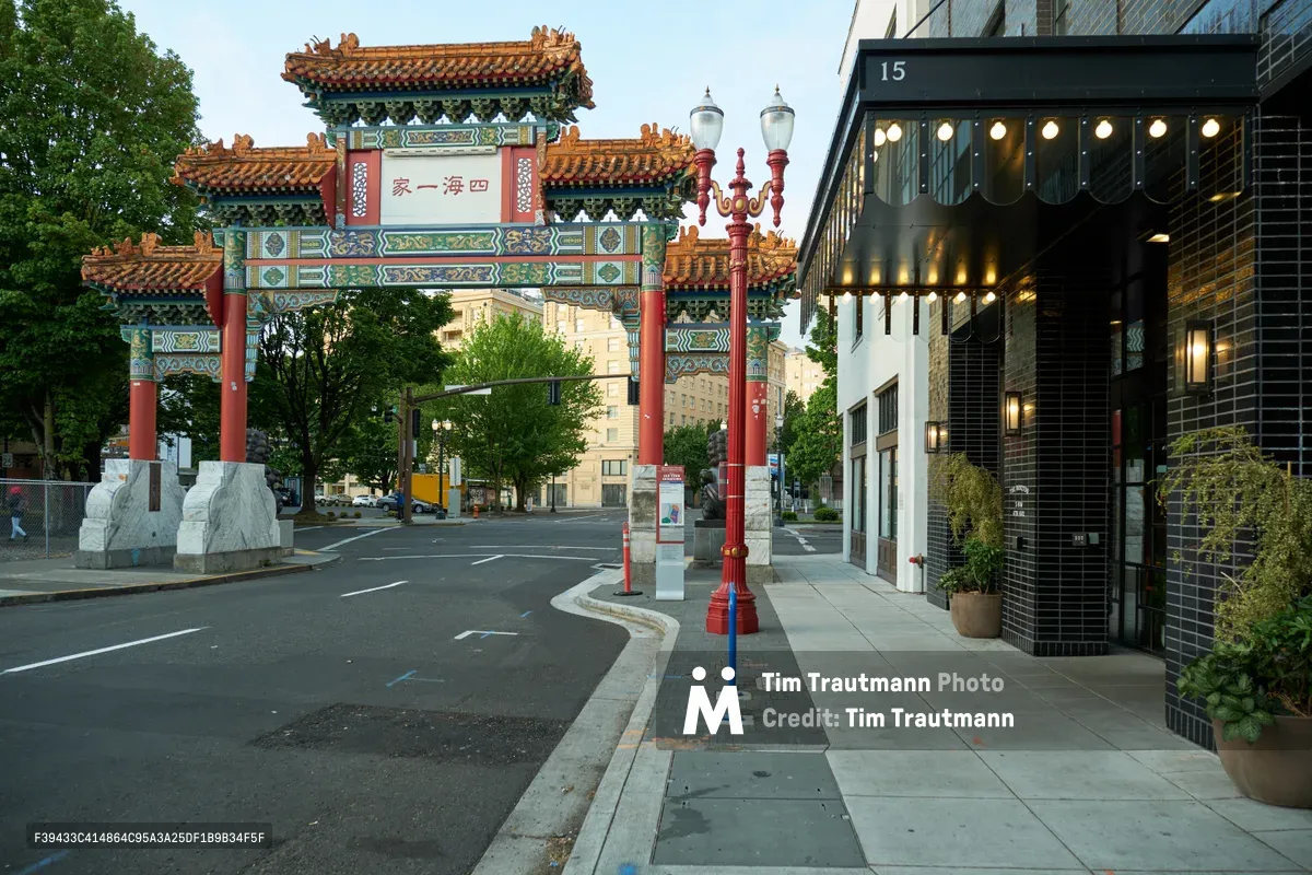 A vibrant Chinese ceremonial archway stands as the entrance to Portland's historic Chinatown district, its ornate red pillars and golden-tiled rooflines creating a striking contrast against the contemporary urban landscape. The early morning light bathes the empty street in a warm glow, while modern buildings including The Hoxton Hotel frame the traditional architecture. String lights from a nearby establishment add a gentle sparkle to the quiet intersection, capturing the seamless blend of cultural heritage and modern city life.