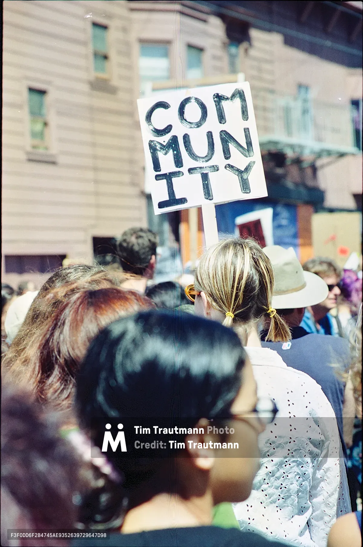 A handmade protest sign reading 'COMMUNITY' in bold black lettering pierces through a crowd of demonstrators gathered in San Francisco's Mission District. The scene captures the raw energy of grassroots activism as residents rally against displacement, with the weathered urban architecture of Bryant Street creating a backdrop of fading authenticity. Blonde pigtails and diverse heads bob through the frame, their faces partially obscured, emphasizing the collective nature of this neighborhood resistance movement.