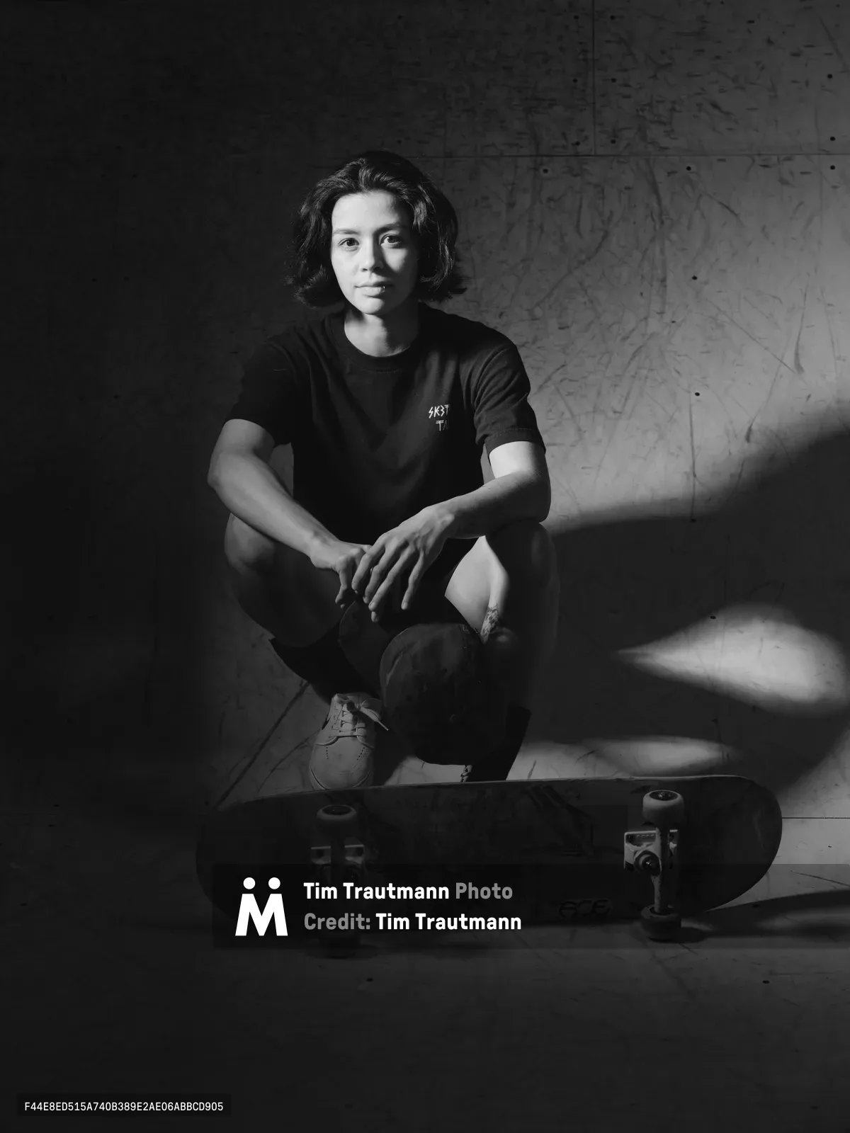 Pro skateboarder Christiana Means sits in contemplative repose against weathered concrete walls at Stronger Skatepark in Milwaukie, Oregon. The monochromatic portrait captures her relaxed posture with arms draped over bent knees, her skateboard positioned prominently in the foreground. Dramatic lighting carves shadows across the textured surfaces, creating an intimate study of athletic grace in an urban sanctuary.