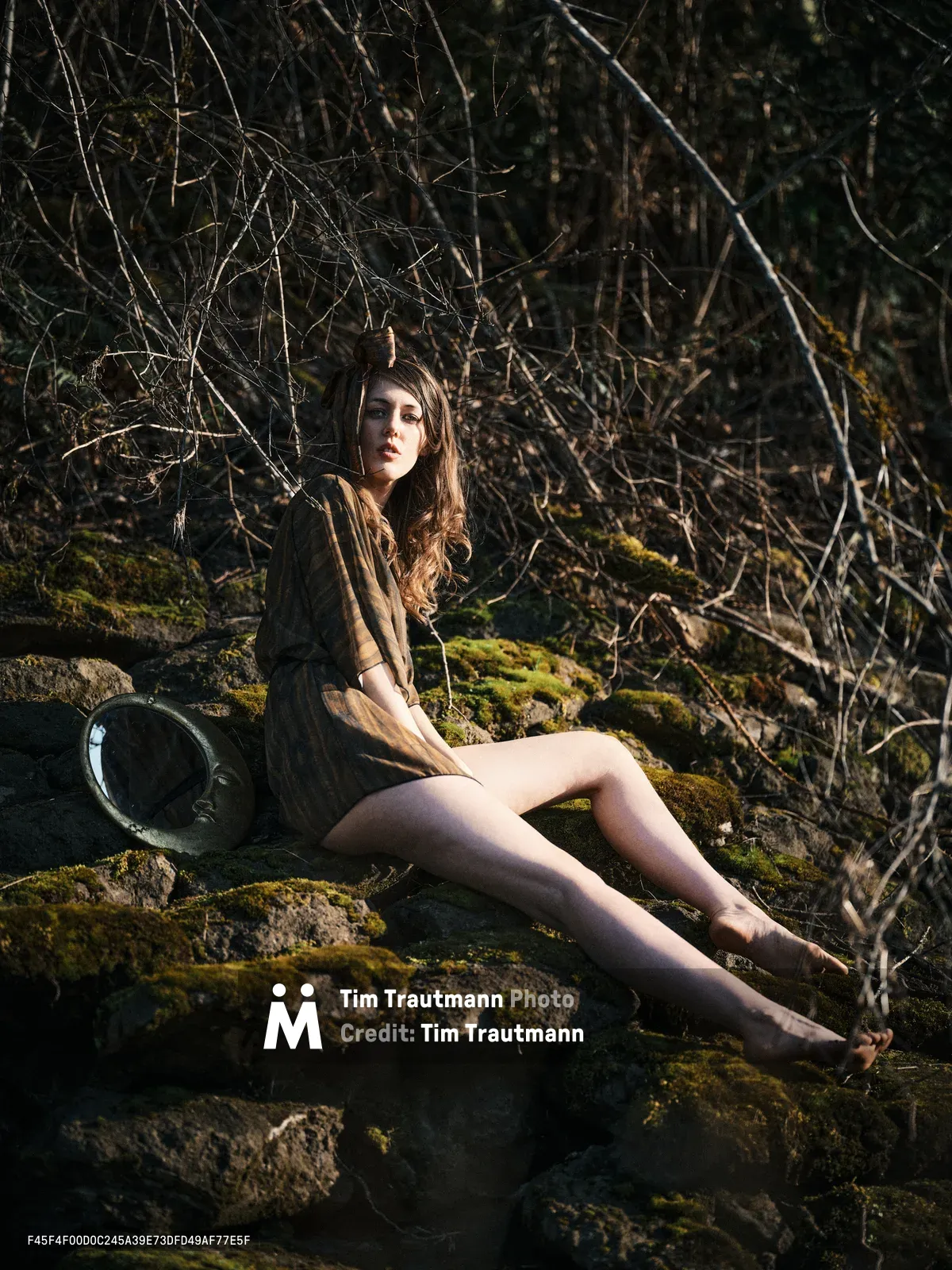 A young woman with long flowing hair sits barefoot on moss-covered rocks along Oregon's Floodplain Trail, bathed in golden hour light that filters through the dense woodland behind her. She wears an earth-toned jacket and holds what appears to be a hat, gazing contemplatively into the distance. The composition captures the intimate relationship between human presence and the ancient, moss-draped stone formations characteristic of the Pacific Northwest's riverbed environments.