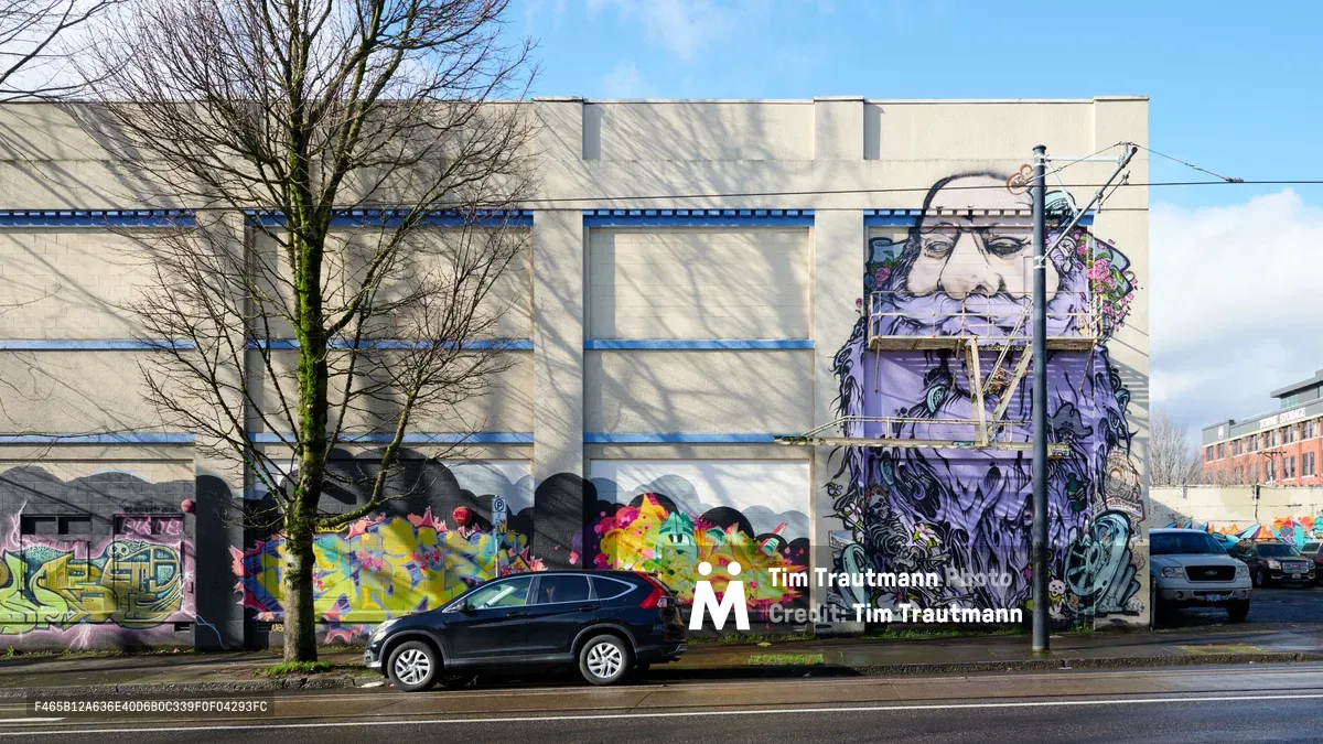 Vibrant murals cascade across the concrete facade of the former Salvation Army warehouse in Portland's Central Eastside district, where colorful graffiti-style artwork mingles with a striking portrait of an elderly bearded figure. The Portland Street Art Alliance's commissioned pieces transform the industrial building into an outdoor gallery, with bare winter trees casting intricate shadows across the wall while vehicles pass along Southeast Ash Street. The juxtaposition of raw urban architecture and expressive street art captures the evolving character of this historically industrial neighborhood.