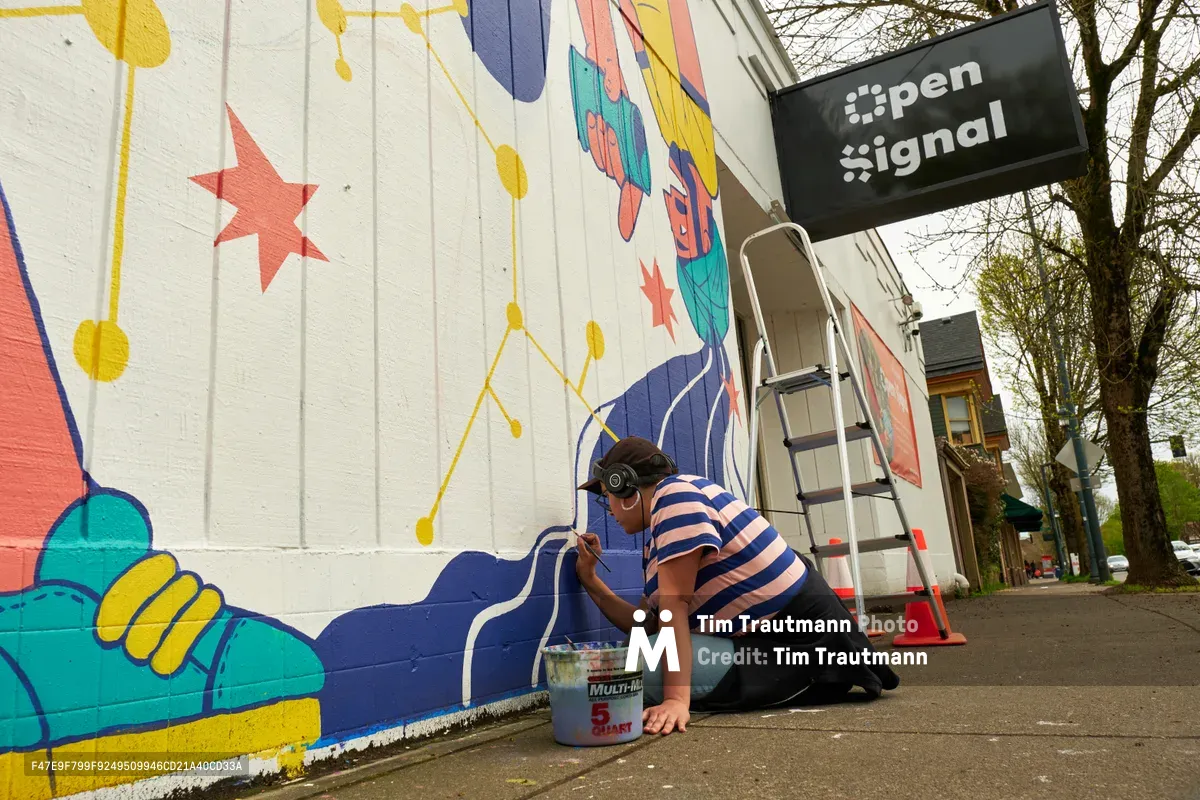 Mexican-American artist Maria Rodriguez, working under the name Sparkykneecap, kneels beside her vibrant mural titled 'Let's Talk' on the exterior wall of Open Signal in Portland's Eliot neighborhood. Wearing a navy and white striped shirt and headphones, she carefully applies paint to the lower portion of her colorful composition featuring abstract figures, stars, and geometric shapes in coral, teal, and golden yellow. A paint bucket sits nearby on the sidewalk as spring light filters through bare tree branches overhead.