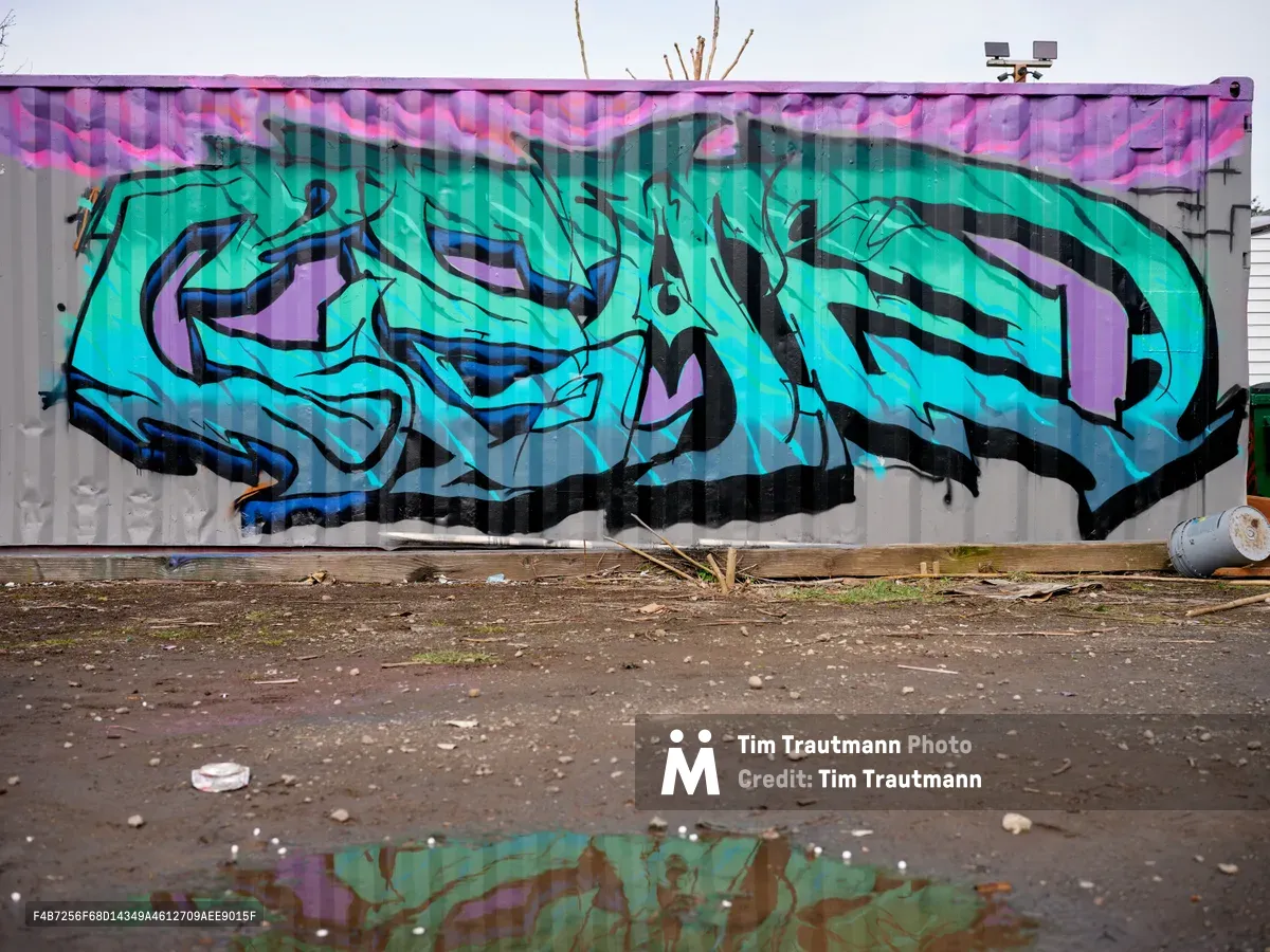 A vibrant turquoise and purple graffiti piece sprawls across an industrial wall in Northeast Portland, Oregon, its flowing wildstyle letters creating an abstract composition of interlocking forms. The artwork's electric colors reflect in a rain puddle on the weathered asphalt below, where scattered debris and weeds emerge through cracks in the pavement. Overcast Pacific Northwest skies cast even lighting across the scene, emphasizing the contrast between urban decay and artistic expression in this post-industrial landscape.