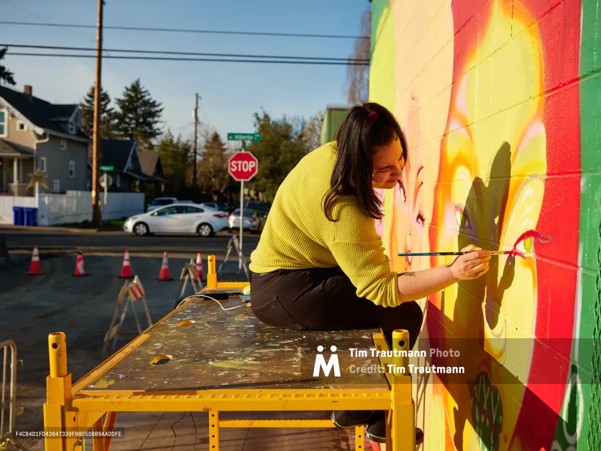 Multidisciplinary artist Villatose meticulously applies brushstrokes to her mural 'Jardín Cubana,' depicting three female figures in vibrant colors on Northeast Alberta Street in Portland's celebrated arts district. Golden hour light bathes the scene as she works from a yellow scaffolding platform, her concentrated expression captured against the vivid spectrum of colors cascading across the wall. The residential neighborhood backdrop, complete with power lines and a stop sign, frames this moment of creative transformation in one of Portland's most dynamic cultural corridors.