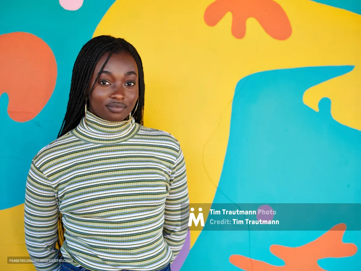 Naomi Likayi stands confidently before her completed abstract mural in Portland's Alberta Arts District, her dark braids framing her face as she wears a striped turtleneck. The dynamic backdrop features flowing organic shapes in brilliant turquoise, golden yellow, coral orange, and soft pink that seem to dance around her figure. The composition captures both the artist's quiet pride and the exuberant energy of her colorful creation, embodying the creative spirit of Northeast Portland's vibrant arts community.