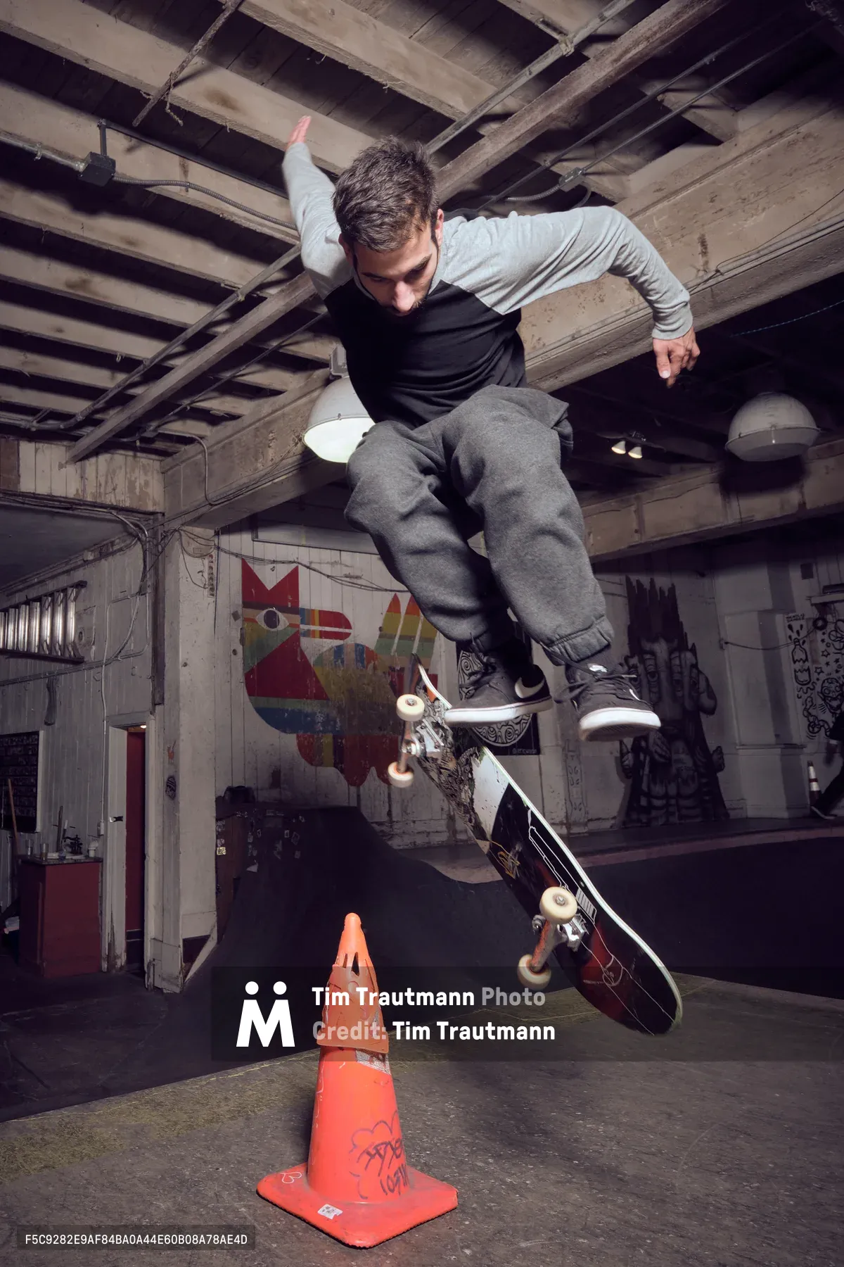 A young skateboarder launches into a gravity-defying ollie over an orange traffic cone in the raw, industrial interior of Commonwealth Skateboarding in Portland's Buckman neighborhood. The weathered wooden ceiling beams and graffiti-tagged concrete walls create a gritty urban canvas, while dramatic lighting captures the precise moment of aerial suspension. His focused expression and athletic form demonstrate the technical mastery required for this fundamental skateboarding trick.