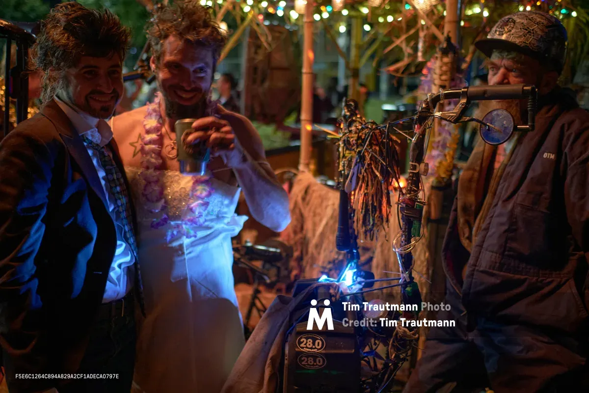 Three men gather around a whimsical bicycle contraption adorned with twinkling lights and decorative tassels during Portland's Drop Out Prom Ride in Colonel Summers Community Garden. The central figure demonstrates the bike's electric illumination system while his companions look on with delight, their faces bathed in the ethereal blue glow. Overhead string lights create a magical canopy, transforming the garden space into an enchanted evening playground for Pedalpalooza's most unconventional celebration.