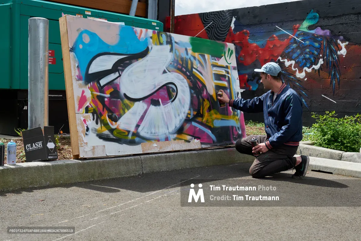 Galen Malcolm kneels beside his vibrant spray-painted canvas during a street art demonstration in Portland's Clay Creative district. The artist, wearing a navy jacket and white cap, adds finishing touches to his colorful graffiti piece featuring bold lettering and dynamic swirls in blues, pinks, and yellows. Behind him, a weathered wall displays established murals, while concrete steps and urban textures frame this intimate moment of artistic creation in the Hosford-Abernethy neighborhood.
