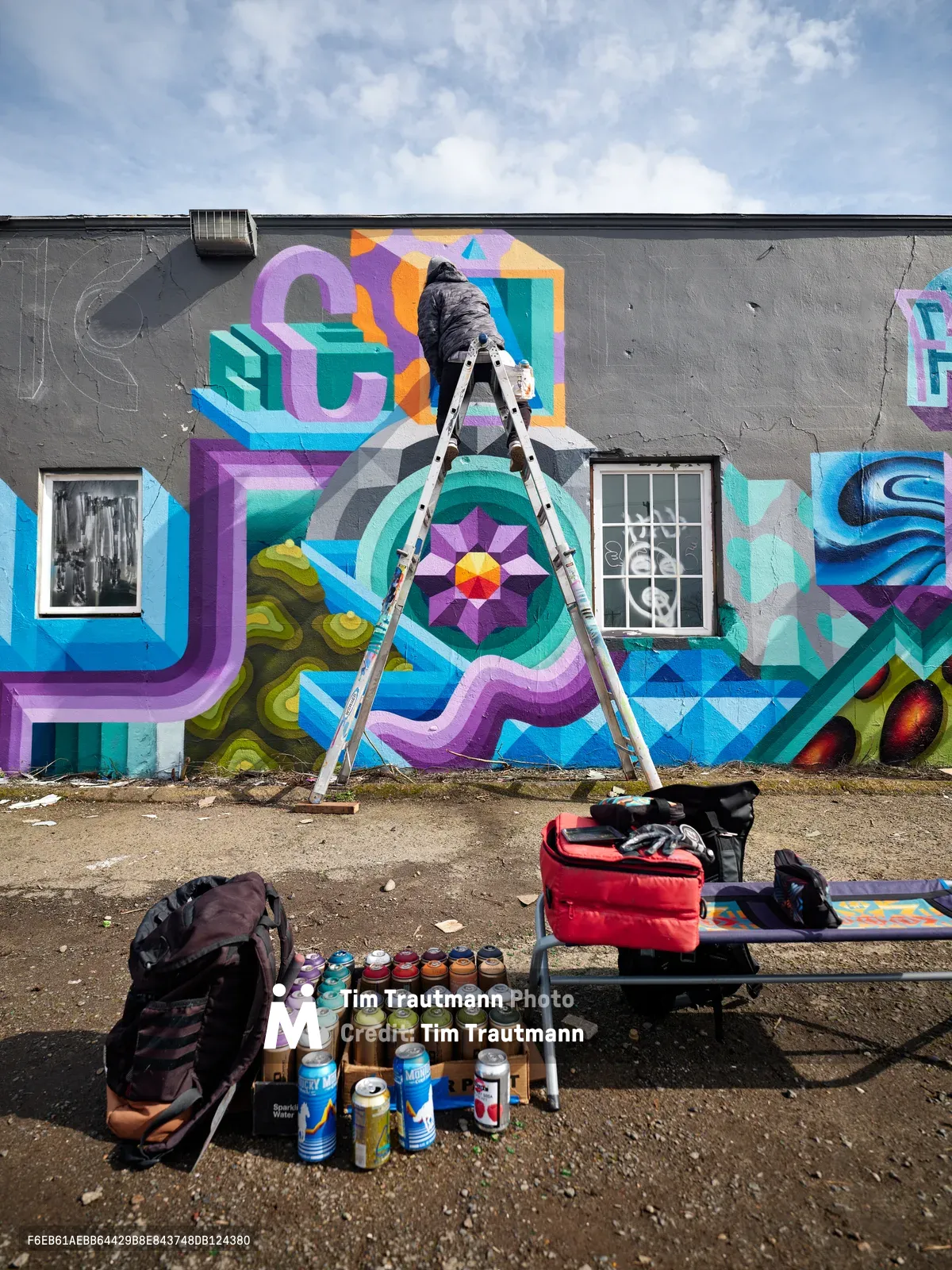 A bundled figure in dark clothing balances on an aluminum ladder, spray painting a dynamic geometric mural across the weathered gray facade of a Northeast Portland building. The kaleidoscopic artwork explodes with interlocking shapes in purple, teal, orange, and electric blue, featuring dimensional letters and a radiating mandala-like centerpiece. At ground level, an arsenal of spray paint cans stands organized in neat rows alongside the artist's equipment, creating a tableau of urban creativity beneath Oregon's characteristic overcast sky.