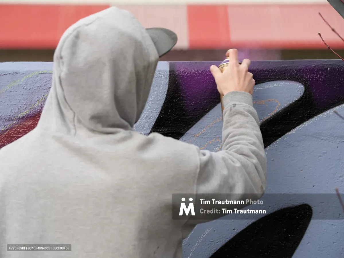 Travis Fields, known as Campographic, works intently on a vibrant mural at the Clay Creative Building in southeast Portland's Hosford-Abernethy neighborhood. Wearing a gray hooded sweatshirt, the artist reaches toward bold purple and black geometric forms painted on the concrete wall. The composition captures the focused concentration of street art creation, with warm coral-pink tones from adjacent surfaces creating atmospheric depth against the cool wall colors.