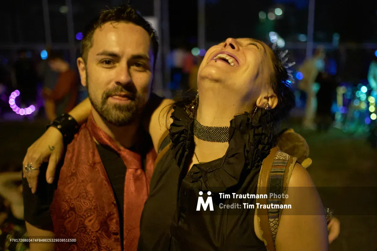 A bearded man in an ornate rust-colored vest wraps his arm around a woman throwing her head back in unrestrained laughter at Portland's Drop Out Prom Ride during Pedalpalooza 2019. The woman's black ruffled dress and choker necklace catch the ambient street lighting as colorful bokeh orbs from the night festival create a dreamy backdrop. Their intimate moment of pure joy unfolds against the blurred carnival atmosphere of Colonel Summers Community Garden, where Portland's alternative cycling celebration transforms the urban landscape into a playground of connection and revelry.