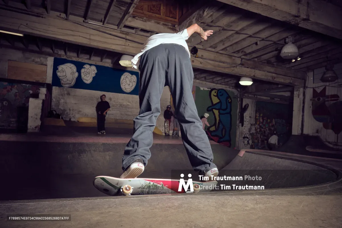 A skateboarder navigates the curved concrete bowl at Commonwealth Skateboarding in Portland's Buckman neighborhood, his baggy grey pants and white shirt creating dynamic lines against the weathered walls. The industrial space hums with underground energy, its exposed wooden beams and graffiti-adorned walls bearing witness to countless sessions. Onlookers gather in the shadows while pendant lights cast a warm glow over this temple of skateboard culture.