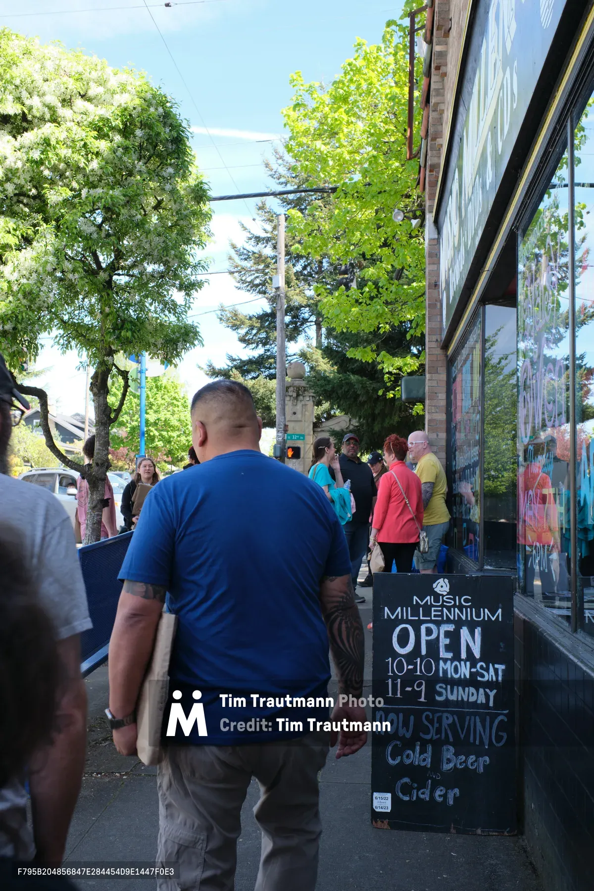A diverse crowd of music enthusiasts gathers on the tree-lined sidewalk outside Music Millennium on East Burnside Street in Portland's Kerns neighborhood. The iconic independent record store's storefront displays vibrant album artwork in its windows, while a prominent chalkboard sign announces operating hours and cold beverages available. Dappled afternoon sunlight filters through the urban canopy, casting a warm glow over the patient queue of customers waiting to enter this beloved musical institution.