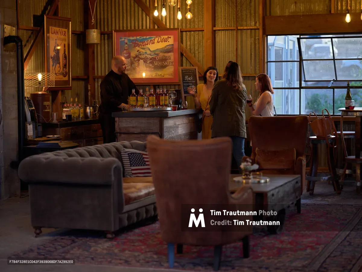 The warmly lit interior of the cocktail bar at Abbey Road Farm in Carlton, Oregon. A bartender in a black shirt stands behind a rustic wooden bar stocked with bottles of whiskey, as three women chat and enjoy drinks. Vintage whiskey advertising posters, including ones for Uam Var Scotch Whisky and Dunard Dew Old Irish Whiskey, hang on corrugated metal walls. Edison bulbs dangle from the ceiling, and the eclectic lounge area in the foreground features tufted velvet sofas, worn leather armchairs, and a Persian rug. A large industrial window lets in soft evening light on the right.