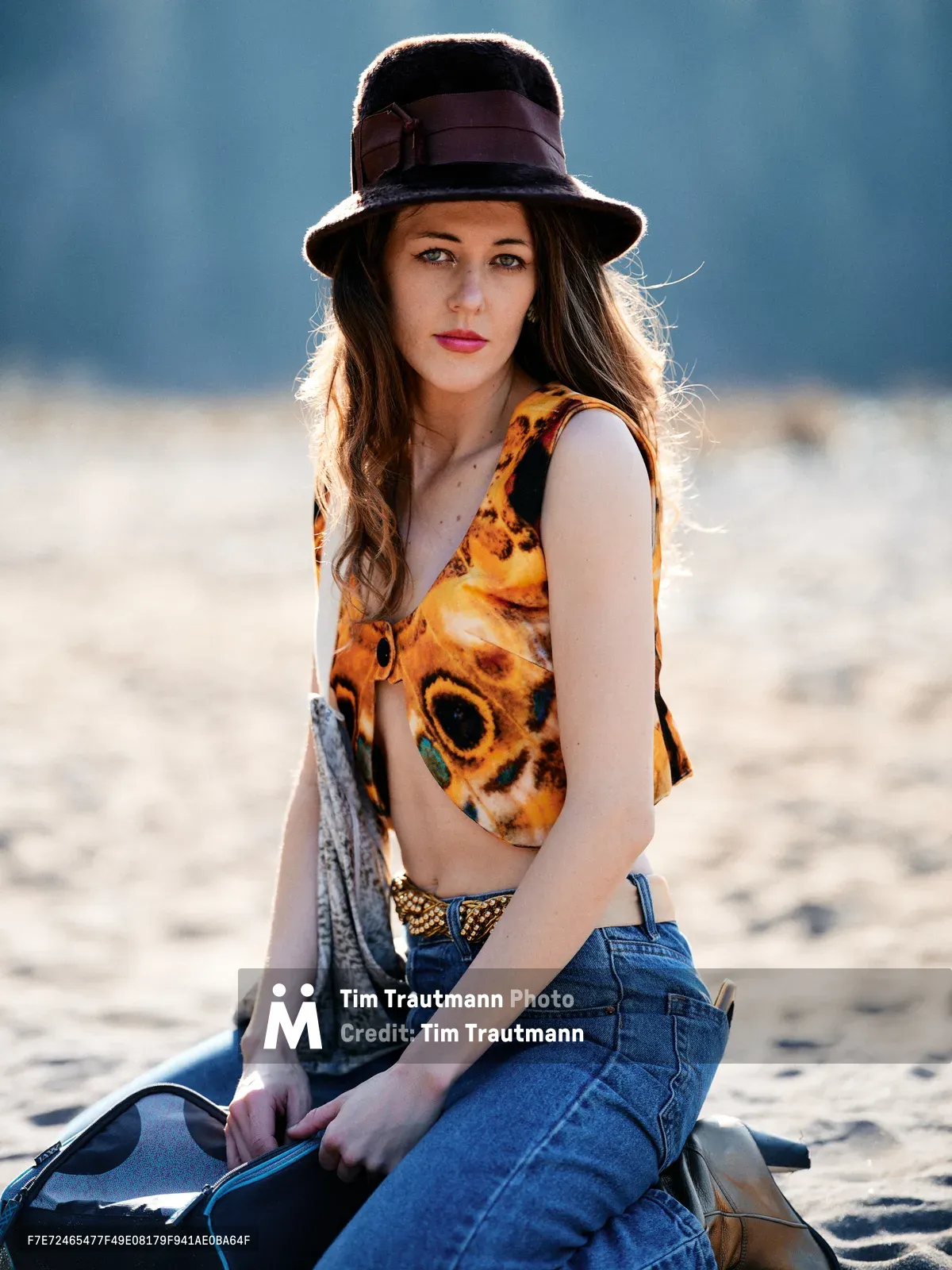 A young woman with long brown hair sits contemplatively on sandy riverbed terrain along the Floodplain Trail near the Sandy River in Multnomah County. Golden hour sunlight creates a warm rim light around her dark felt hat with burgundy band, while she wears a vibrant butterfly-print tank top and high-waisted denim. The soft, diffused lighting and muted blue-gray background of distant hills create an intimate, bohemian atmosphere in this Pacific Northwest setting.